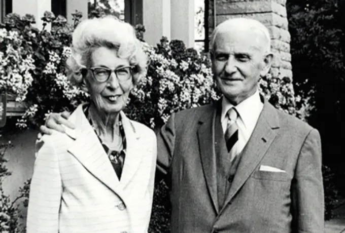 Ein historisches Schwarz-Weiß-Foto der Grunder von der Fondation, Ernest Lory und Thérèse Henchoz, das lächelnd nebeneinander steht. Die Frau trägt eine helle Jacke und eine Brille, während der Mann einen dunklen Anzug mit Krawatte trägt. Hinter ihnen ist ein Gebäude mit Fenstern, umgeben von blühenden Sträuchern, zu erkennen, was auf ein gepflegtes Haus oder einen Garten hindeutet.