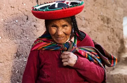 Ein Foto einer lächelnden älteren Frau in traditioneller andiner Kleidung. Sie trägt einen leuchtend roten Poncho, mehrfarbige Textilschärpen über der Schulter und einen charakteristischen, breitkrempigen Hut, der mit einem bunten Band verziert ist. Die warmen Farben ihrer Kleidung heben sich deutlich von der erdfarbenen Wand im Hintergrund ab.