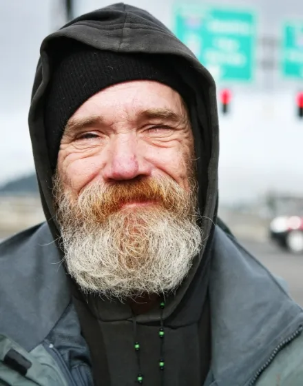 Porträt eines älteren Mannes mit einem freundlichen Lächeln. Er hat einen vollen grauen Bart, Tränen in den Augen und trägt eine schwarze Kapuze sowie eine schwarze Jacke mit einem sichtbaren grünen Muster am Kragen. Im Hintergrund sind unscharfe Straßenschilder und Verkehr, was darauf hindeutet, dass das Foto im städtischen Außenbereich aufgenommen wurde.