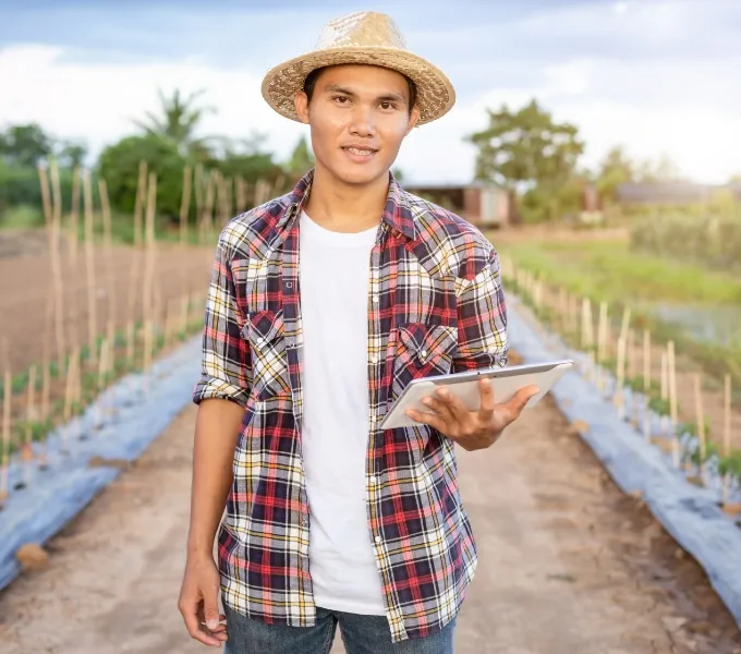 Ein junger Mann steht in einem landwirtschaftlichen Feld, er trägt einen Strohhut und ein kariertes Hemd über einem weißen T-Shirt. Er hält ein Tablet in seiner Hand und blickt in die Kamera, mit einem leichten Lächeln. Im Hintergrund sieht man die gepflegten Beete des Feldes und den bewölkten Himmel eines frühen Abends oder Morgens.