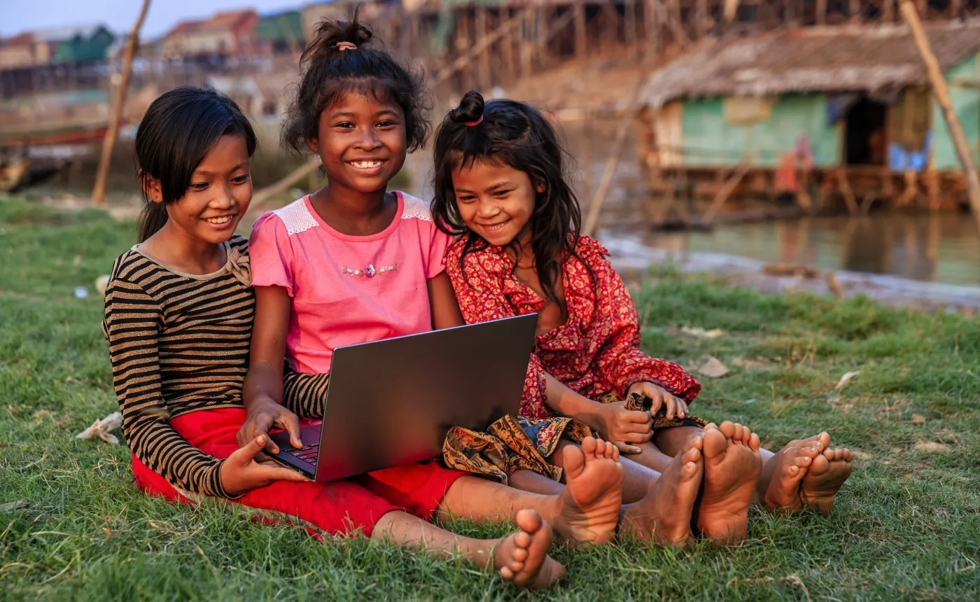 Drei junge Mädchen sitzen gemeinsam auf einer Wiese und lächeln, während sie auf einen Laptop schauen, den sie gemeinsam halten. Die Mädchen sind barfuß und tragen bunte Kleidung; eine trägt ein gestreiftes Oberteil und eine rote Hose, eine andere ein pinkes Shirt und die dritte ein rotes Musterkleid. Im Hintergrund sieht man auf Stelzen gebaute Häuser über einem Gewässer, was auf eine ländliche Wasserdorf-Szene hindeutet.