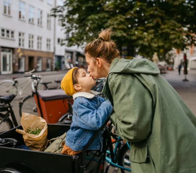 Eine Frau in einer grünen Jacke gibt einem kleinen Kind, das eine gelbe Mütze und eine blaue Jeansjacke trägt, einen Kuss auf den Mund. Sie befinden sich neben einem Lastenfahrrad mit einem Einkaufsbeutel darin, was auf eine urbane Umgebung hinweist. Im Hintergrund sind unscharfe Gebäude und geparkte Fahrräder zu sehen.